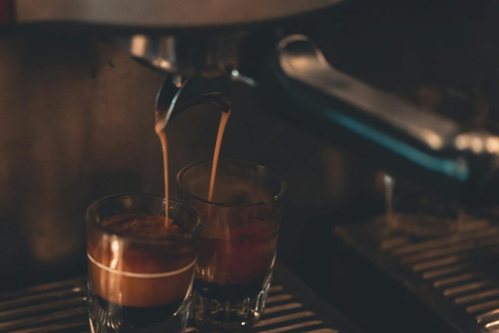Brew Your Burn. Close-up view of hot espresso pouring into two glasses, creating a rich coffee aroma indoors.