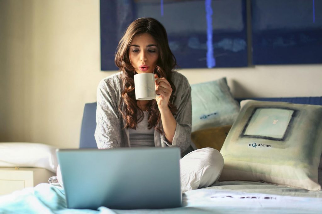coffee origins, A woman enjoying coffee while working from home in a cozy bedroom setting with a laptop.